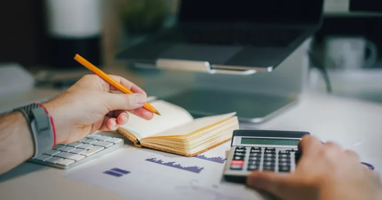 Pessoa com calculadora e caderno fazendo contas em uma mesa de trabalho