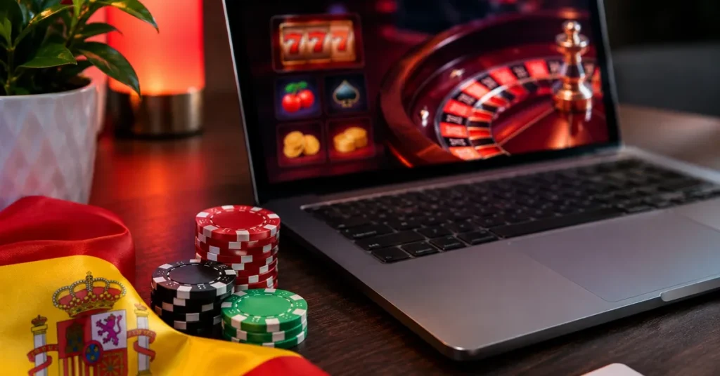 Fichas de casino, una bandera de España y un portátil con una ruleta y una tragaperras online en pantalla
